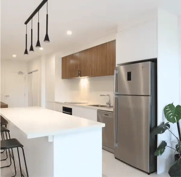 Modern kitchen with white countertops, wooden upper cabinets, stainless steel refrigerator, and black pendant lights.