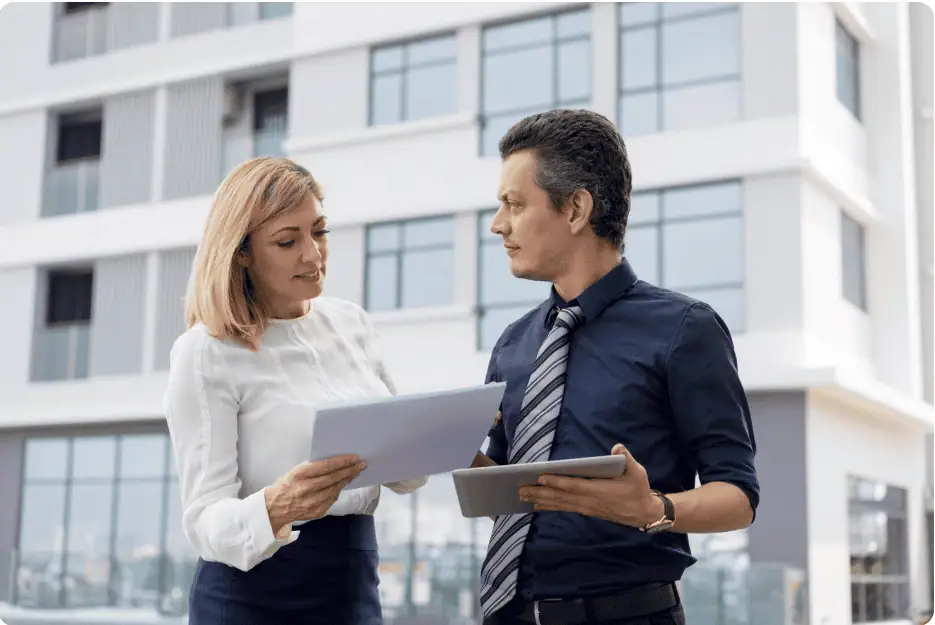 Two business professionals, a woman and a man, discussing documents outside a modern office building.